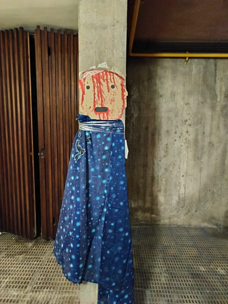 A fake creepy guy with a bloody cardboard face and a blue, broken, starry curtain (i think it's a curtain jhfsd) tied to a column.