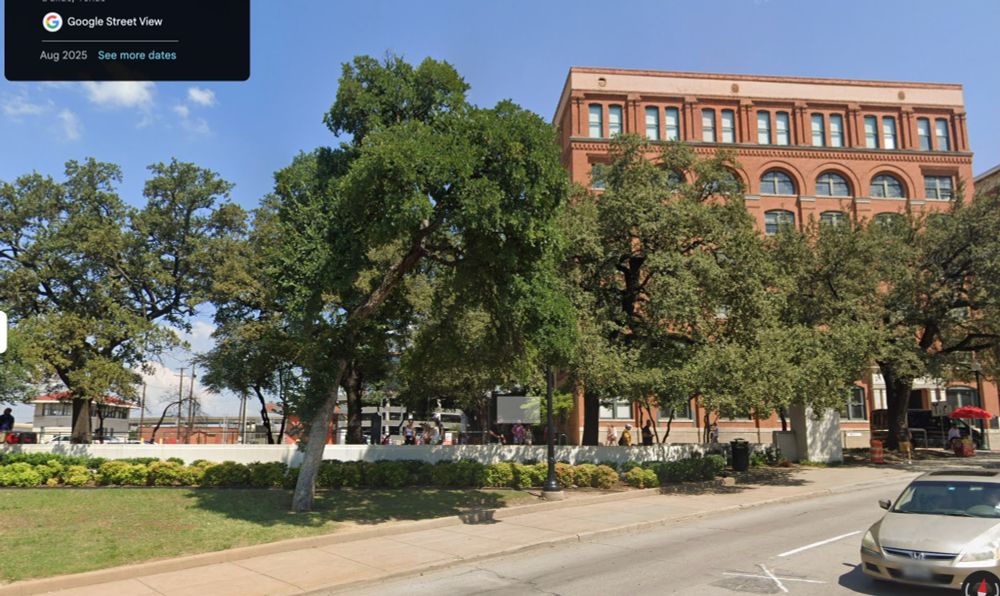 The Texas School Book Depository on the right side picture, facing north on Elm St with the grassy knoll to the left. A low white fence extends between the grass and the parking lot behind it, at perhaps waist height for passing pedestrians.