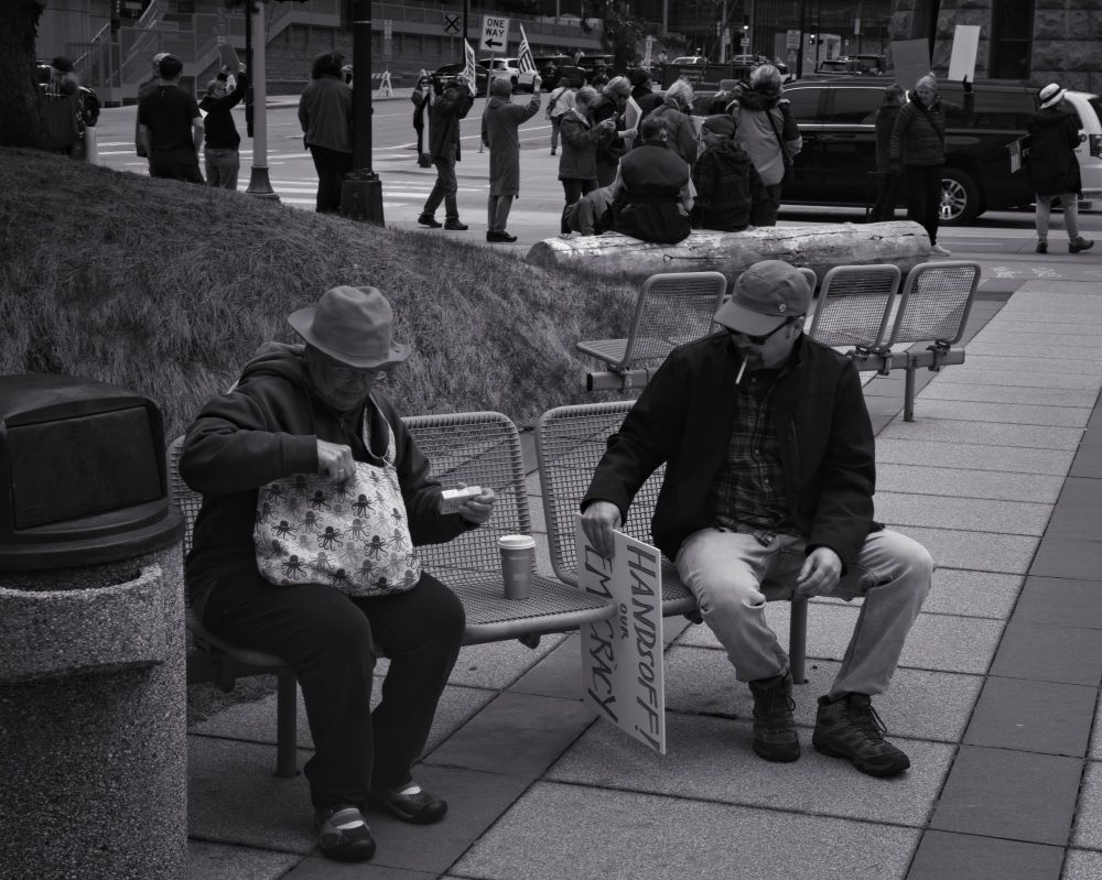 A monochrome photo of two people sitting on a metal bench in a city setting. The person on the right has a cigarette in their mouth, and is setting down a protest sign. The person on the left is putting a pack of cigarettes into their bag. In the background can be seen a crowd of protesters with a multitude of signs.