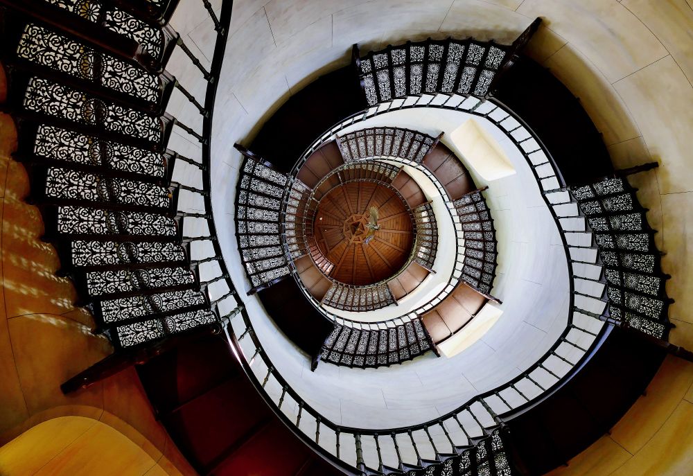 Blick nach oben
eine schwarze Wendeltreppe aus Metall mit Mustern in den Treppenstufen zirkelt sich an hellen Wänden nach oben