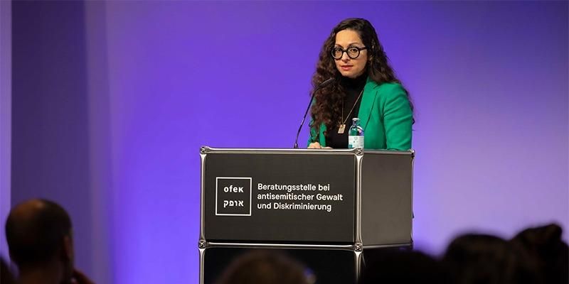 Marina Chernivsky auf einem Podium der ofek Beratungsstelle bei antisemitischer Gewalt und Diskriminierung vor einem violetten Hintergrund. Im Vordergrund sind verschwommen die Köpfe von Zuschauenden zu sehen. Bildrechte: Ruthe Zuntz.
