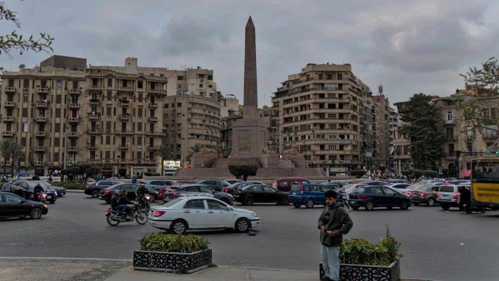 Autos fahren über einen Platz, in der Mitte steht ein Obelisk. Im Hintergrund mehrstöckige Wohnhäuser.