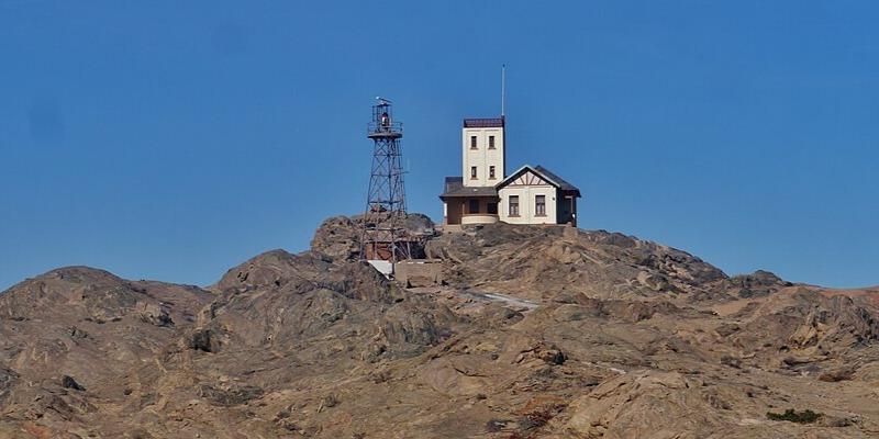 Leuchtsignal auf der Haifischinsel, Lüderitz, Region Karas, Namibia. Man sieht ein Haus mit einem erhöhten Anbau auf einem felsigen Hügel. Links daneben und leicht versetzt darunter steht ein Leuchtsignalturm aus Metallstreben. Die Landschaft ist sehr bergig. Im Hintergrund ein strahlend blauer Himmel ohne Wolken. Foto: Zairon.