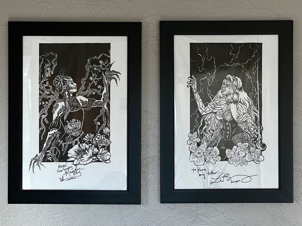 A photo of two linocut prints hanging on a wall in black frames. On the left is Laudna and on the right is Imogen Temult, both from critical role. They sort of face each other, with their hands reaching up towards each other. Marisha Ray signed the Laudna print with “Kenz! Fun Scary!” And Laura Bailey signed the Imogen print with “to Kenz- my tether…”