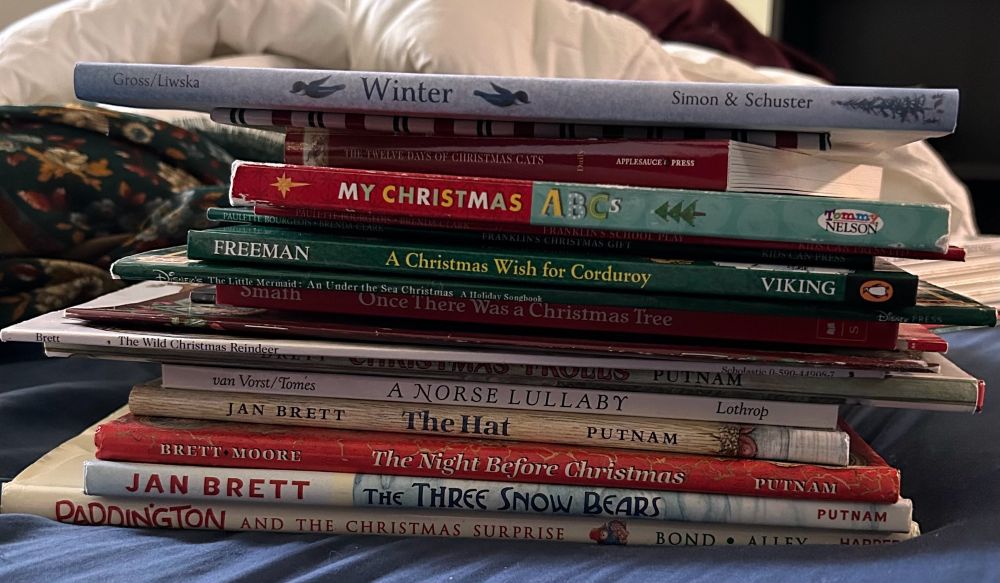 A photo of a stack of children’s holiday books resting on a bed, including titles by Jan Brett, Paddington Bear, Corduroy, and a couple board books, topped by a book about the Winter Solstice.