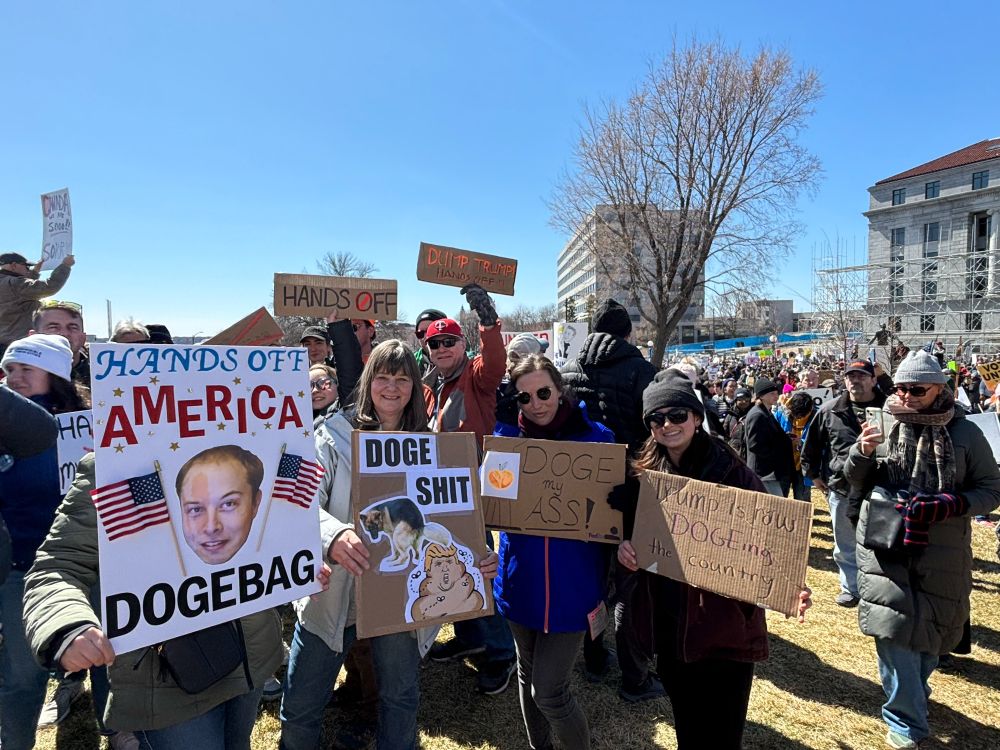 People with signs prominent signs saying America DOGE Bag with image of Elon Musk, DOGE Shit with image of a dog pooping a Trump-poop emoji.