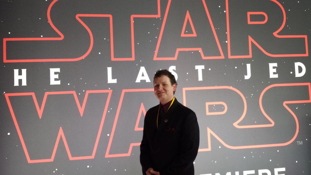 A photo of a man wearing a dark suit with a dark red shirt, a black necktie, a yellow lanyard, and a "Join the Resistance" pin on his lapel. He's standing in front of a wall that has the words "Star Wars: The Last Jedi" on it.