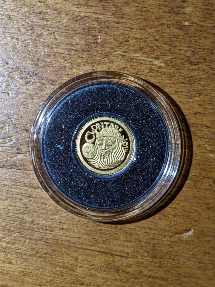 A photo of a small, yellow coin on a wooden desk. The coin is in a protective case and surrounded by black spongy padding. On the visible face of the coin is a stylized face of a man with a flowing beard and a crown. To the left of the man is a circle with "20 EURO" in it. Text spanning in a curve along the coin face's rim from the circle to the other side of the man's head reads "CLONTARF 1014".