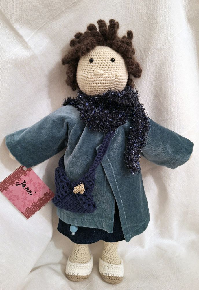 Foto einer Häkelpuppe mit blauem Samtmantel und einem darunter hervorschauendem Jeansrock. Der Mantel hat einen Schalkragen aus blauer Glitzerwolle, dazu kommen weiße Häkelschuhe.  Die Puppe hat braune kurze Locken, alle gehäkelt. Sie trägt eine blaue Häkeltasche mit einem silbernen Anhänger in Form zweier Fausthandschuhe. An der Hand hat sie ein Namensschild, auf dem „Janni“ steht.  