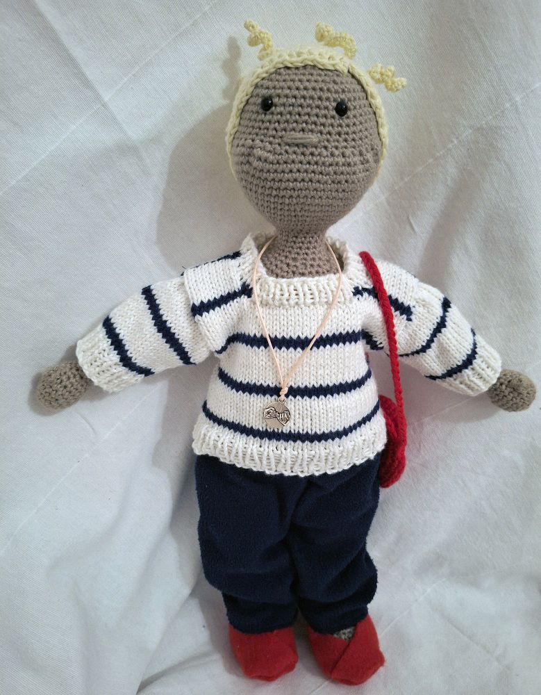 Puppe Jule nun ohne den Regenmantel. Sie trägt Jeans, einen gestrickten weißen Pulli mit maritimen dunkelblauen Streifen. Zudem trägt sie einen silbernen Anhänger um die Hals