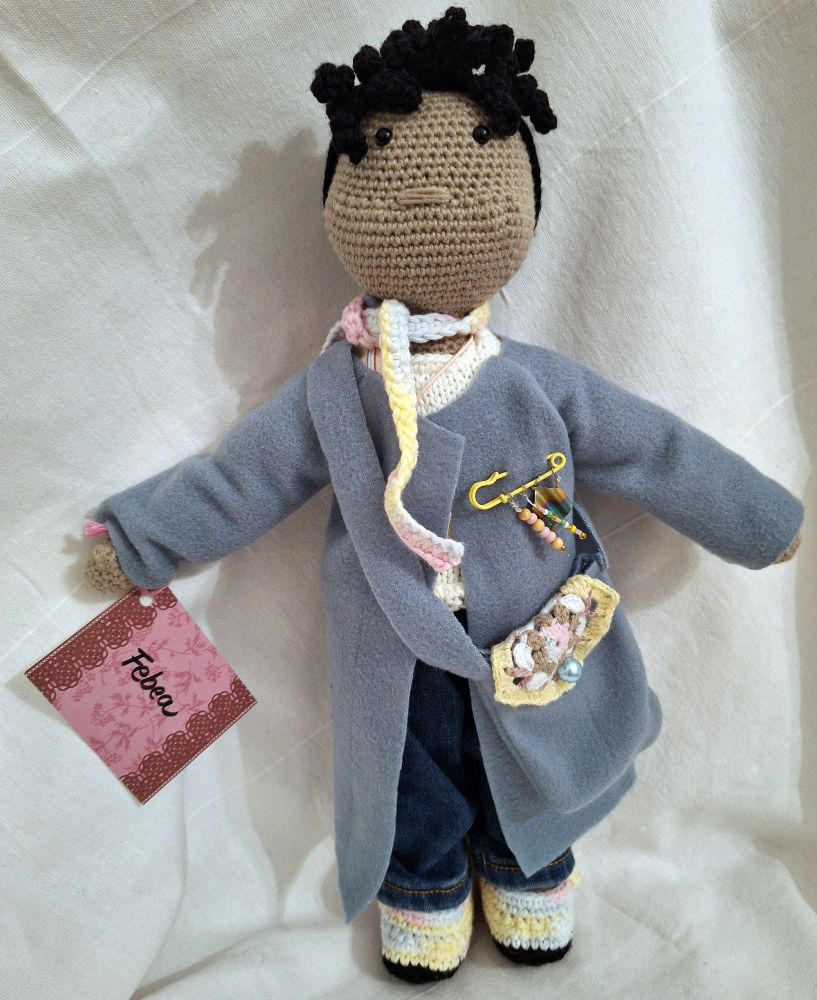 Foto einer Häkelpuppe mit Jeans und hellblauem Fleecemantel. Die schwarzen Haare haben einen lockigen kleinen Pony. Sie trägt einen gehäkelten Schal, eine blaue Fleecetasche mit Granny-Square-Klappe sowie einen gelbe Anstecknadel mit Perlenanhängern daran. Die Schuhe sind ebenfalls gehäkelt und aus derselben pastellmelierten Wolle wie der Schal. Die Puppe ist aus dunkelbeiger Baumwolle gehäkelt und hat schwarze Knopfaugen. An der Hand hat sie ein Namensschild, auf dem „Febea“ steht.