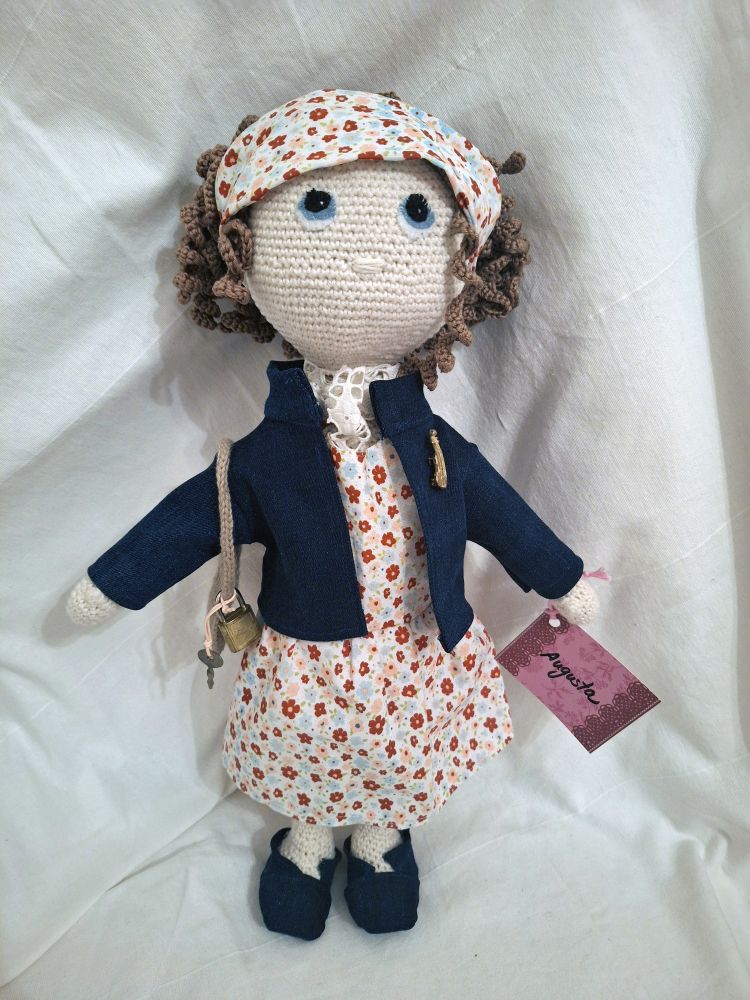 Foto einer Häkelpuppe mit Blümchenkleid und Jeansjacke sowie – schuhen. Sie trägt ein breites Haarband aus dem gleichen Blümchenstoff wie ihr Kleid. An der Jacke hat sie eine kleine braune, ebenfalls gehäkelte Umhängetasche mit einem Schloss und Schlüssel daran. Der Kopf der Puppe ist recht groß, und unter dem Haarband kommen braune, gehäkelte Locken hervor. Die blauen Augen sind aus Fleecestoff aufgenäht. An der Hand hat ein Namensschild, auf dem „Augusta“ steht.