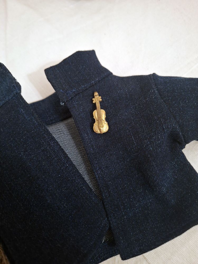 Detailfoto der Jeansjacke von Puppe Augusta mit einer kleinen goldenen Brosche in Geigenform