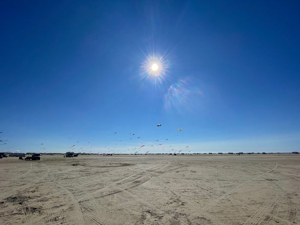 Blauer Himmel im oberen Teil des Bildes. Mittig die strahlende Sonne. Im Himmel tanzen jede Menge Drachen. 
Im unteren Teil flacher Sandstrand. In der Ferne sieht man Autos am Strand.