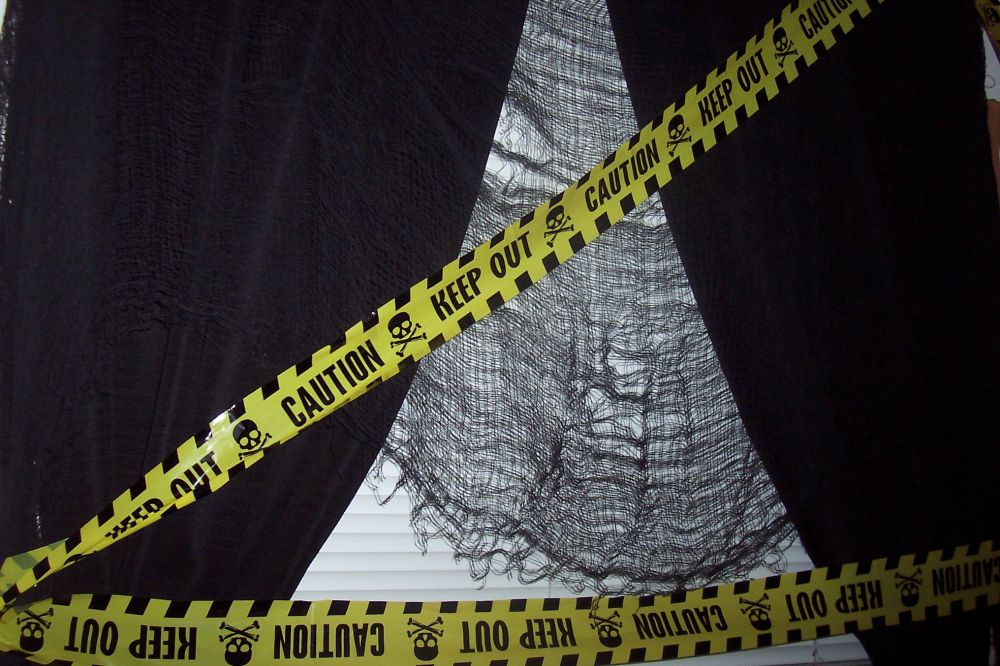 Yellow caution tape with skull-and-crossbones symbols is stretched diagonally across a dark, tattered black cloth over a window with blinds visible behind it.