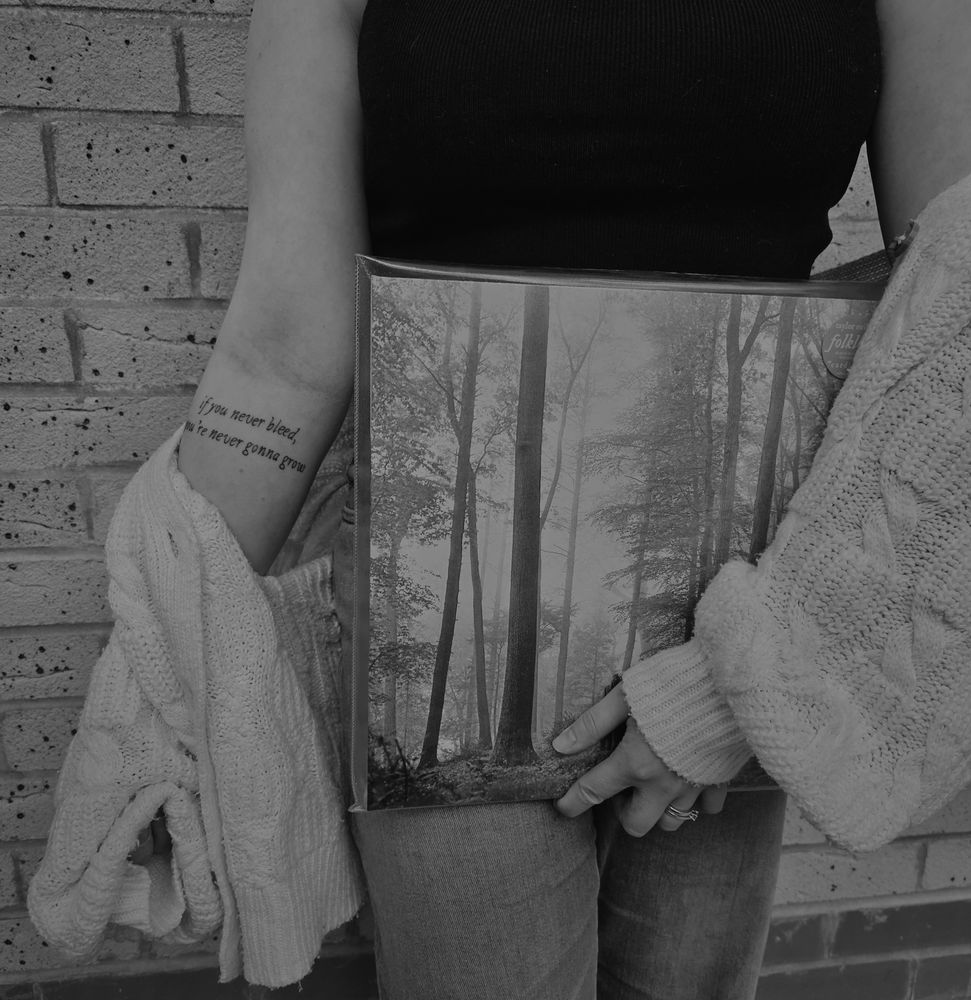 A black and white photo of woman holding a folklore vinyl, with a white cardigan draped down her right arm showing a tattoo that reads “if you never bleed you’re never gonna grow”. The picture cuts off at the shoulders so you cannot see her face