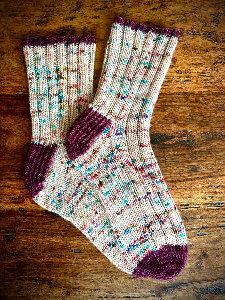A hand knit pair of socks are displayed on a wooden table. The toes, heals and top of cuff are burgundy wine colored in contrast with the off white with multi-colored speckles main body of the sock.