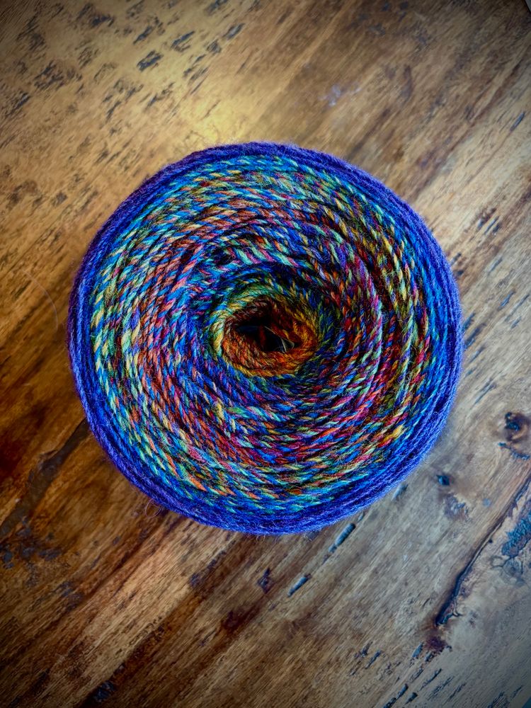 A round cake of handspun yarn is displayed on a wooden background. The yarn transitions colors of the rainbow as it moves from the center outwards.