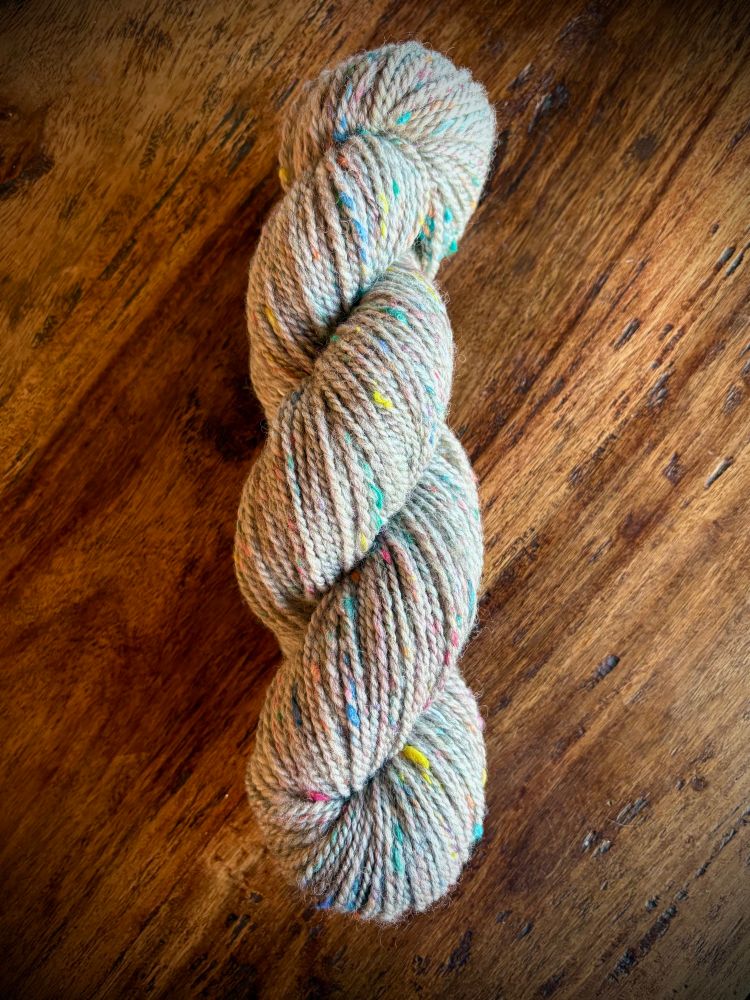 A single skein of grey yarn with tweedy rainbow colored bits of sari silk throughout is displayed on a wooden table.