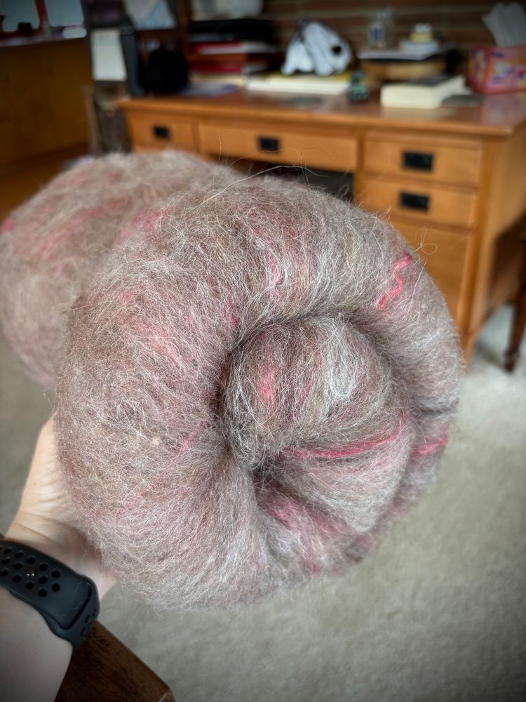 A spiral rolled, wool batt grey in color with streaks of red is held in a woman’s hands. 