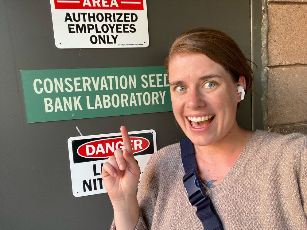 Selfie of a white woman with brown hair smiling with her mouth open standing in front of a door and pointing at a sign reading “Conservation Seed Bank Laboratory”. Other signs on the door read “Danger Liquid Nitrogen” and “Authorized Employees Only”. 