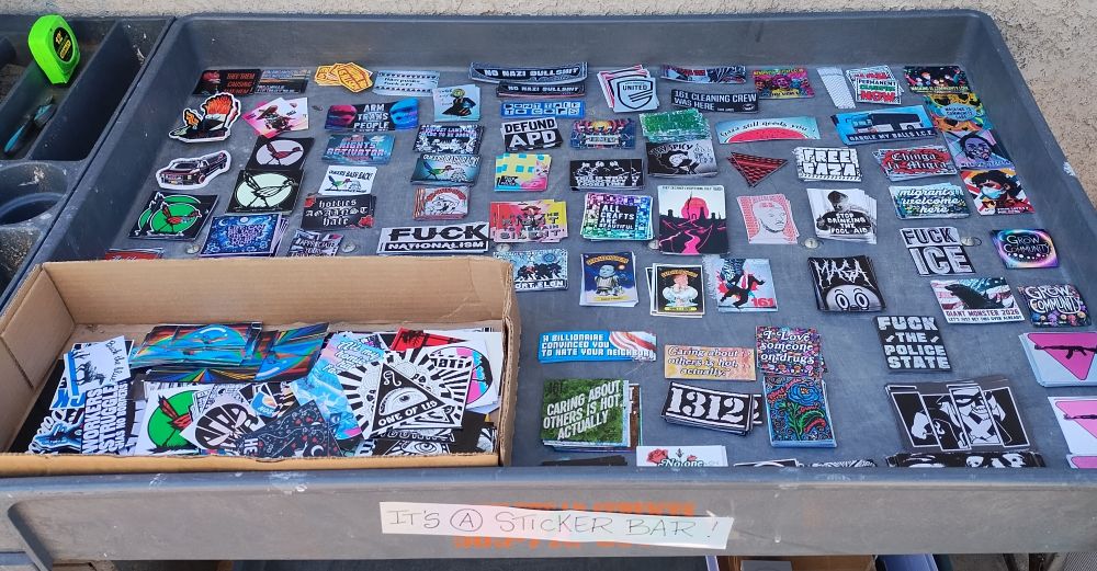 Image shows a table filled with stickers of various left leaning statements, pro Palestine, pro queer, pro trans, pro community, anti cop,anti nationalist, anti maga. You get the idea. 