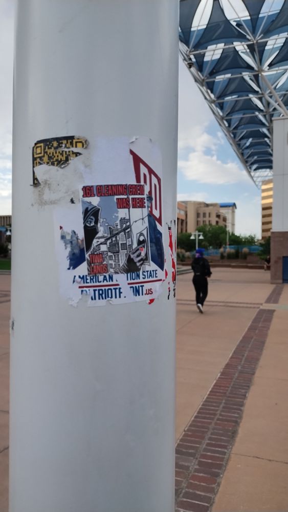 Photo of the torn remains of a racist bullshit sticker covered up with another sticker. The cover up sticker shows a person in black bloc putting an afa sticker on a wall. It reads 161 cleaning crew was here. Tewa land.  It is on a pole at Albuquerque civic plaza. 