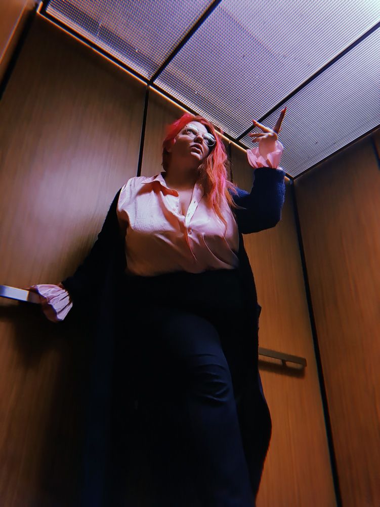 Satine stands above you in an elevator with hand outstretched towards the ceiling. They are wearing a pink billowy shirt and long cloak. Their shirt is unbuttoned to the 2nd button and you can almost see a peak.
