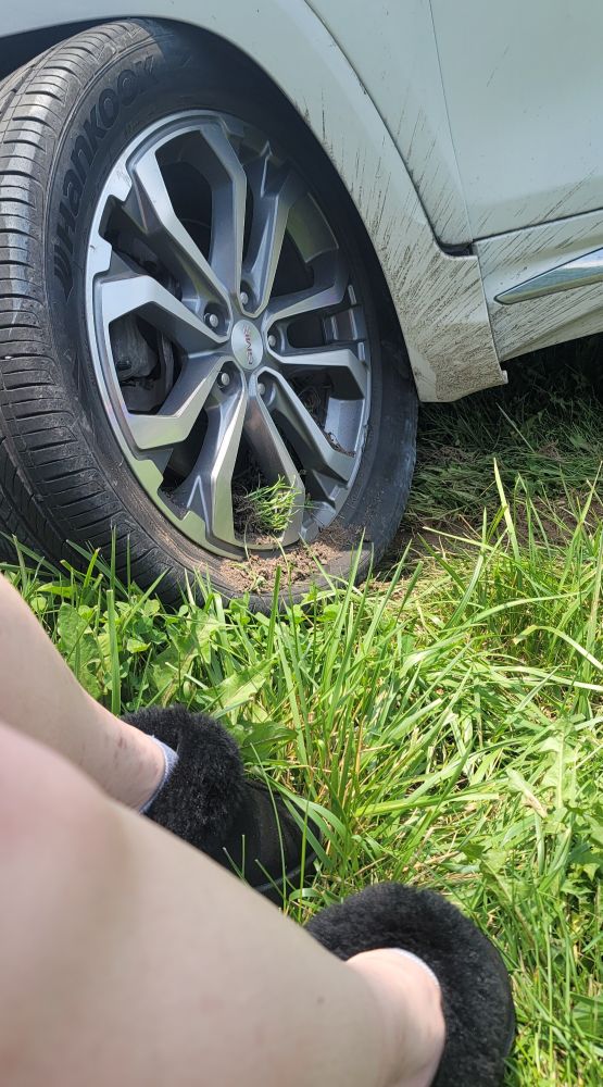a young person's legs and feet wearing fuzzy black slides (shoes) sitting in the grass next to the front end of a car that has been badly damaged in an accident.