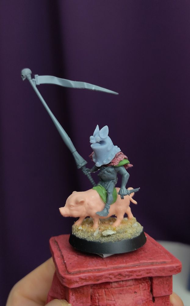 A photo of a miniature, the miniature is a goblin riding a a pig while holding a lance with a skull and pennant on the end and the goblin is holding a shiv in the other hand and is wearing a helmet that looks like a cute bat face.