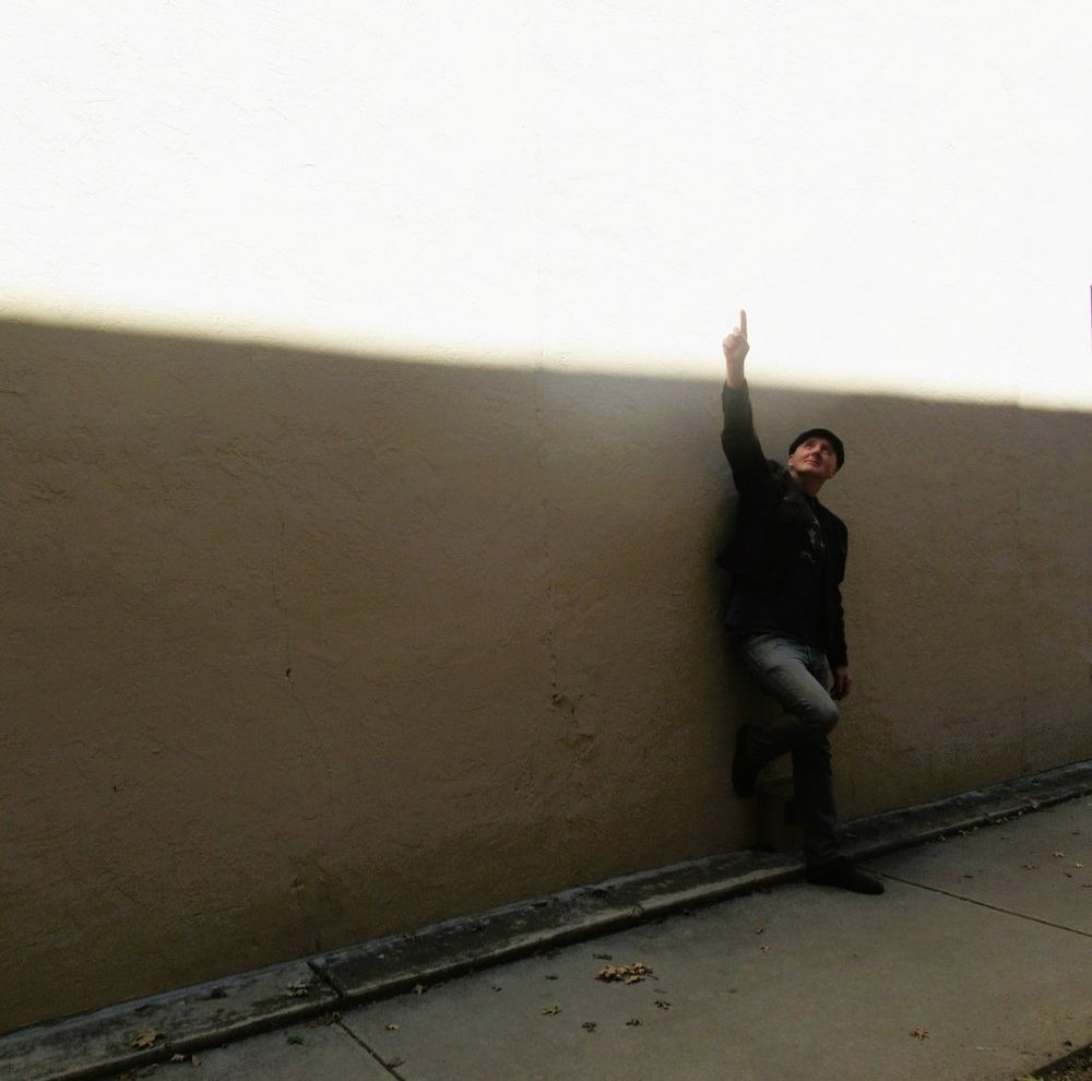 A man stands against a wall where sunlight and shadow are sharply delineated. He points upward to the source of the light.  