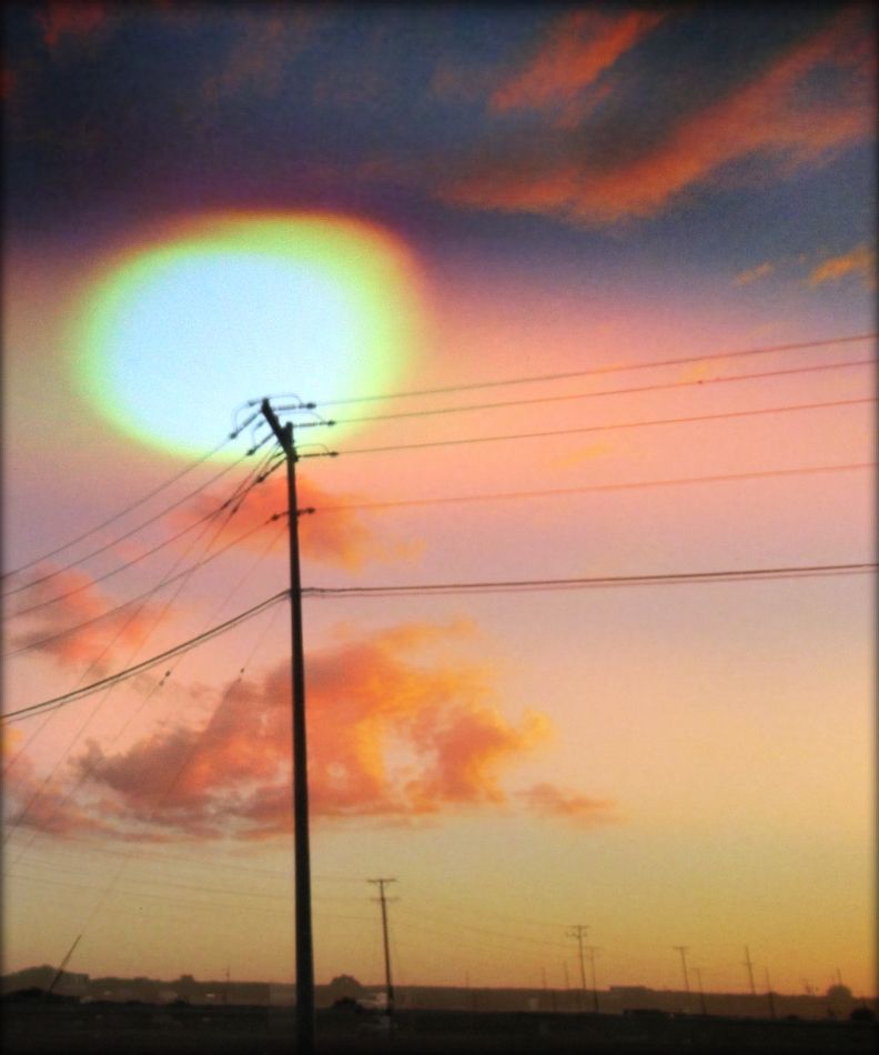 A telephone pole and wires in silhouette against a hazy sun and pink sky with orange clouds in the background.