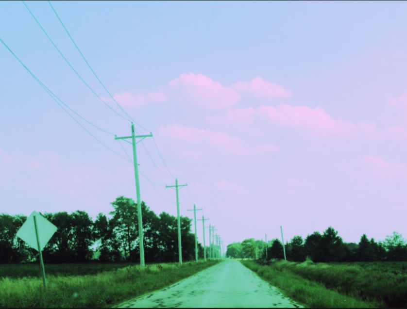 A country road with telephone wires stretching into the distance. There are fields on each side and clouds at the horizon. 