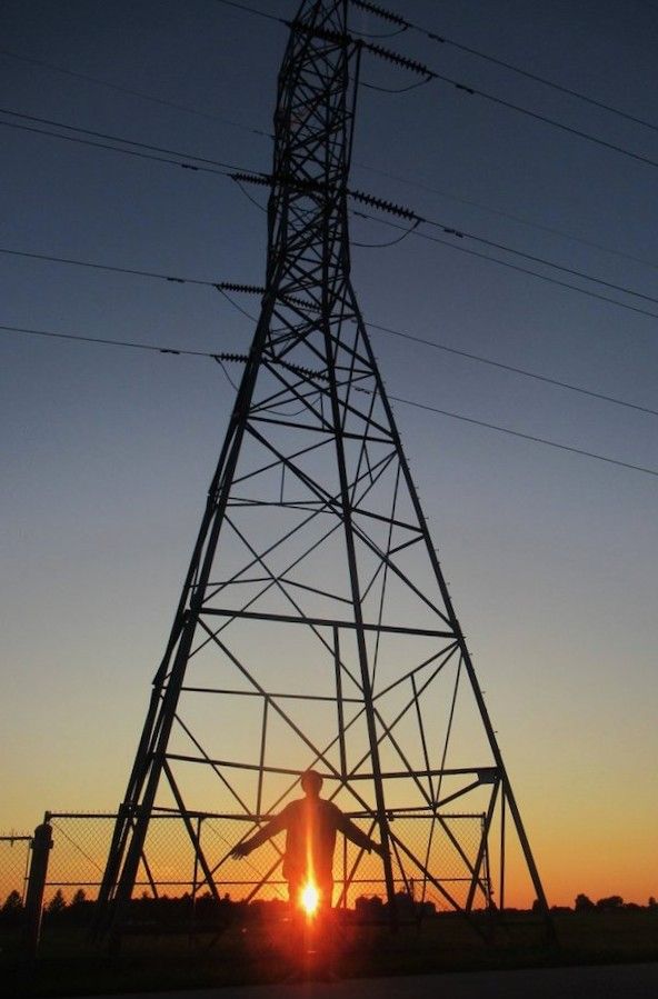 A man, with the light of the setting sun glimmering around him, stands at the base of a pylon with his arms slightly raised. 