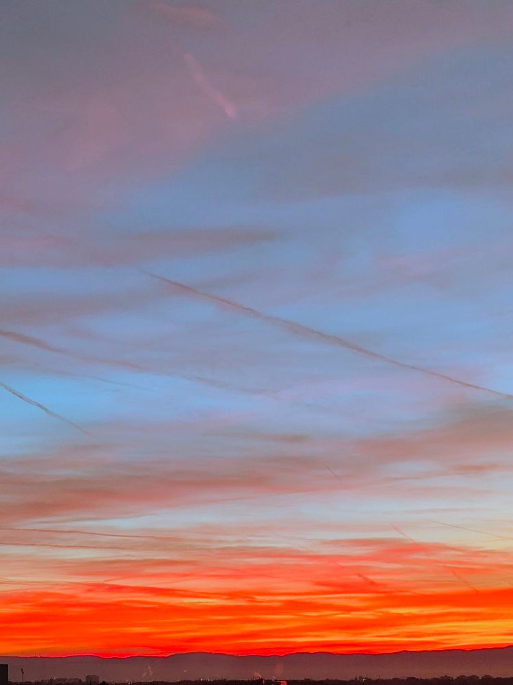 Sonnenaufgang. Der Himmel hat einen Farbverlauf von oben hellblau nach unten kräftig orange. Am Horizont ist eine Gebirgskette zu erahnen. 