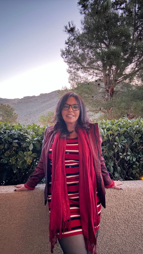 A glamorous woman with dark, voluminous hair poses confidently in a form-fitting burgundy strapless gown, smiling over her shoulder. She wears gold earrings and red lipstick, exuding confidence, class, and sultry charm against a dark, softly lit background.