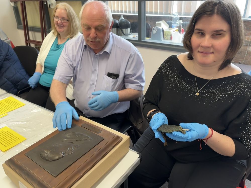 Three individuals wearing blue nitrile gloves. On the right, a woman holds the Great Seal of Edward I (1239–1307), which appears dark with traces of green. In the centre, a man gently touches a bas-relief portrait of Victor Branford (1863–1930) created by Georges Durand.