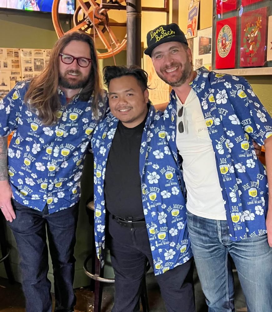 Three men standing closely together inside a bar or restaurant, all wearing matching blue Hawaiian-style shirts that say 'Joe Jost's Long Beach' with white hibiscus flowers and yellow logos. The man in the middle is shorter, with short black hair and a black shirt underneath his open matching shirt. The man on the left has long hair and red glasses. The man on the right has a beard, is wearing a 'Long Beach' baseball cap, and has sunglasses tucked into his shirt. All three are smiling and appear to be enjoying themselves.

