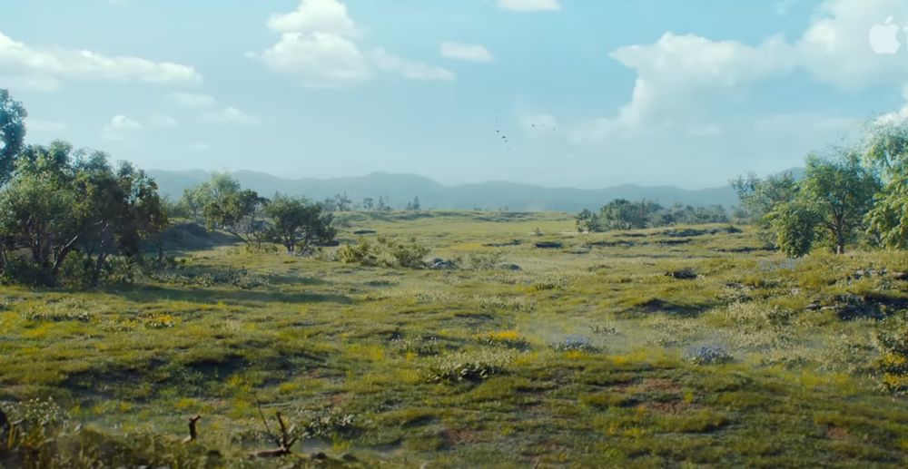 Wide view of a lush, open meadow under a bright blue sky with scattered clouds, surrounded by distant trees and hills, from the Apple TV+ series Silo.