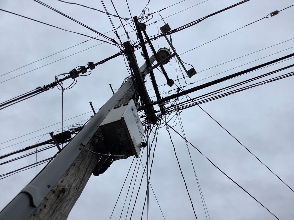 Rise of a wooden power pole through planes of power and communication lines and associated devices, against mottled gray overcast.