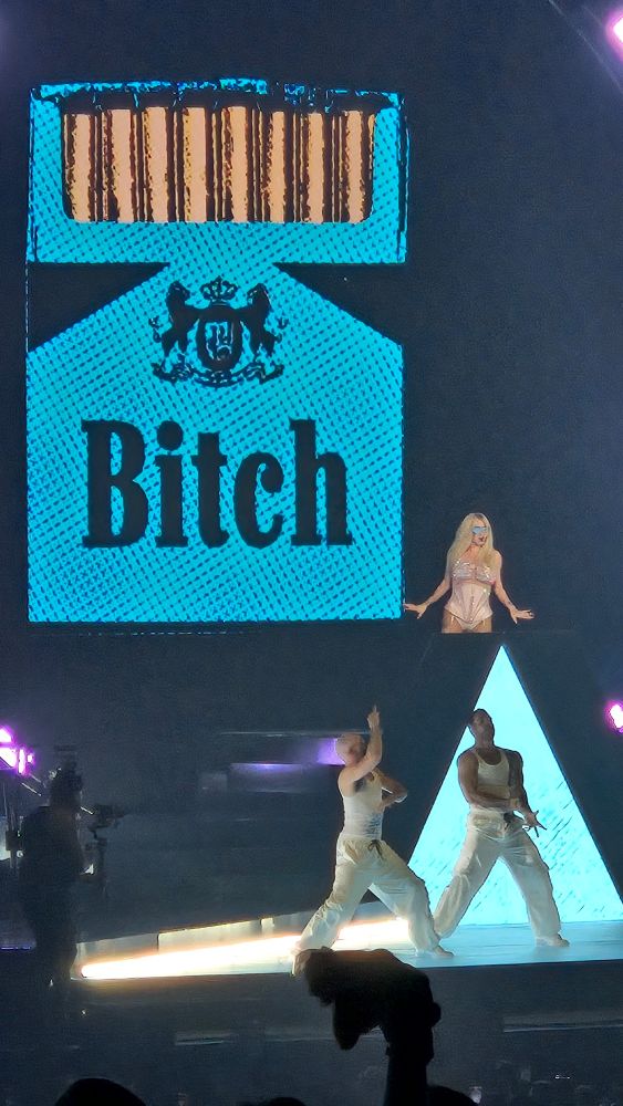 Kesha with backup dancers and a giant sign that says "bitch" on a carton of cigarettes