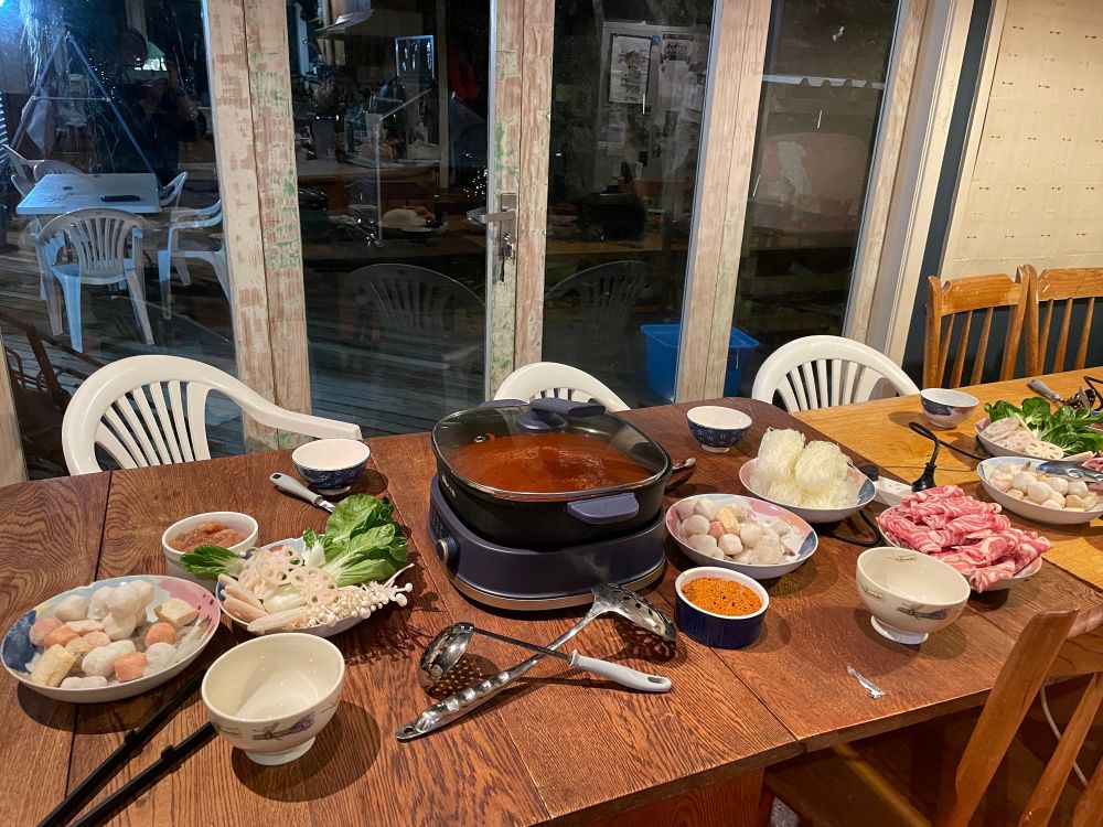 A spread of food on a dining table with an electric pot that has red spicy broth in it. Surrounding it was different plates containing deferent food: assorted fishballs and prawn, thinly sliced lamb, thinly sliced chicken, glass noodles, a plate of vegetables (Pak Choi, enoki mushroom, sliced lotus roots), and some schezuan dipping sauce. Also some slotted spoons and bowls to serve.

