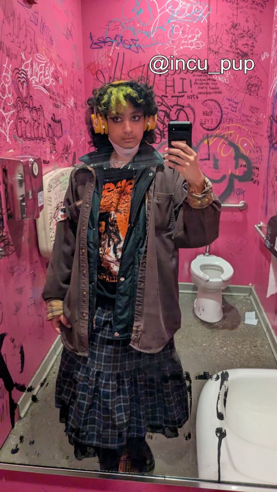Buffy takes a full body mirror selfie in a bright pink bathroom with graffiti on the walls. He has curly black shoulder length hair with a yellow green dyed chunk at the front of his head. He wears yellow over ear headphones, an oversized brown denim jacket, a black shirt with red art on it, and a navy blue plaid skirt.