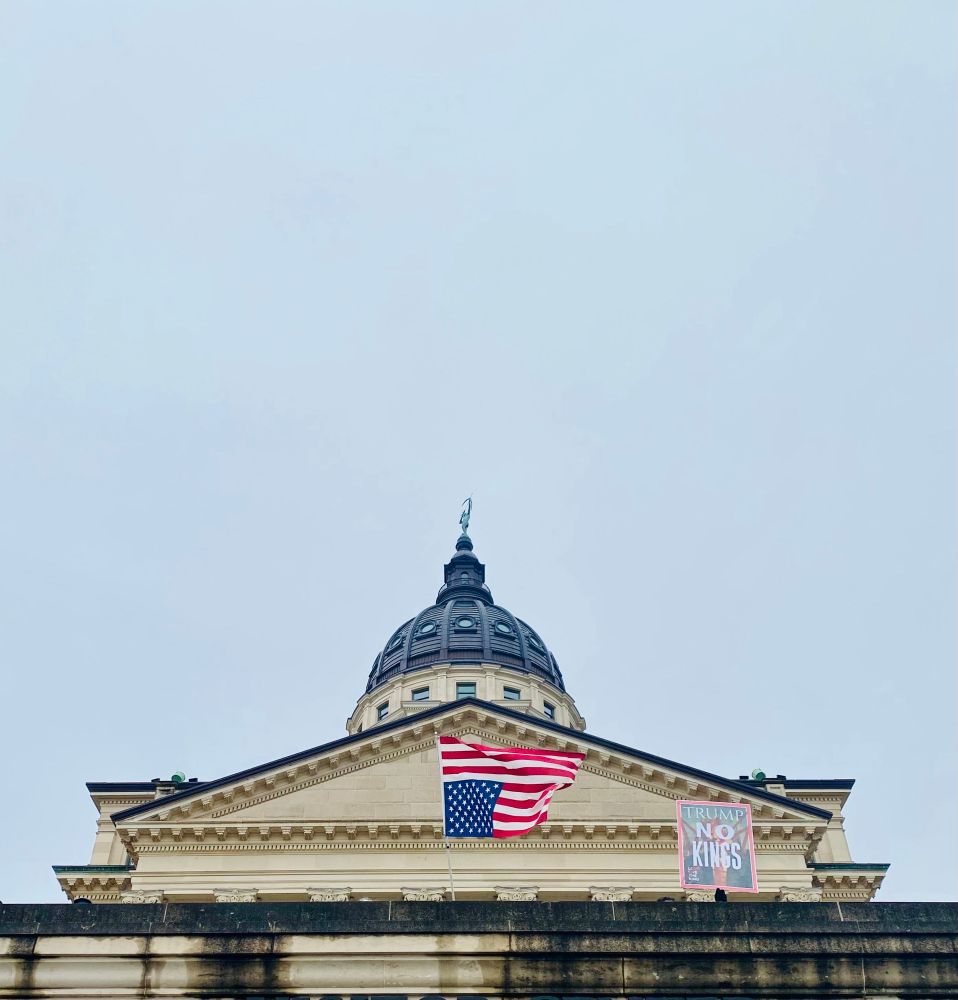 The dome of the Kansas state capitol viewed from a low angle. Protestors hidden from view hold a US flag upside down as a distress signal, and a sign reading "Trump: No Kings."