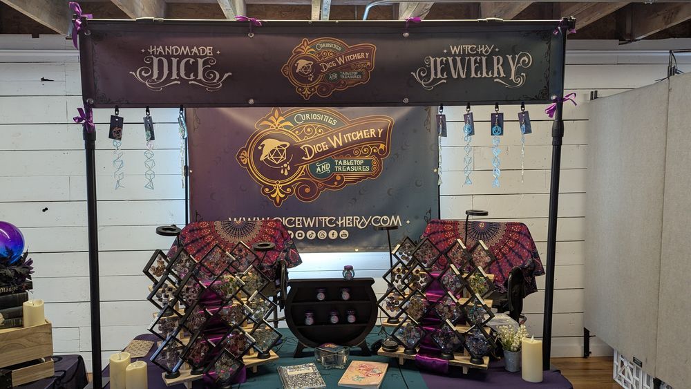 Dice Witchery vendor display. There are dice displays in front. The banners are blue and purple with the Dice Witchery logo and the words "handmade dice" and "witchy jewelry."