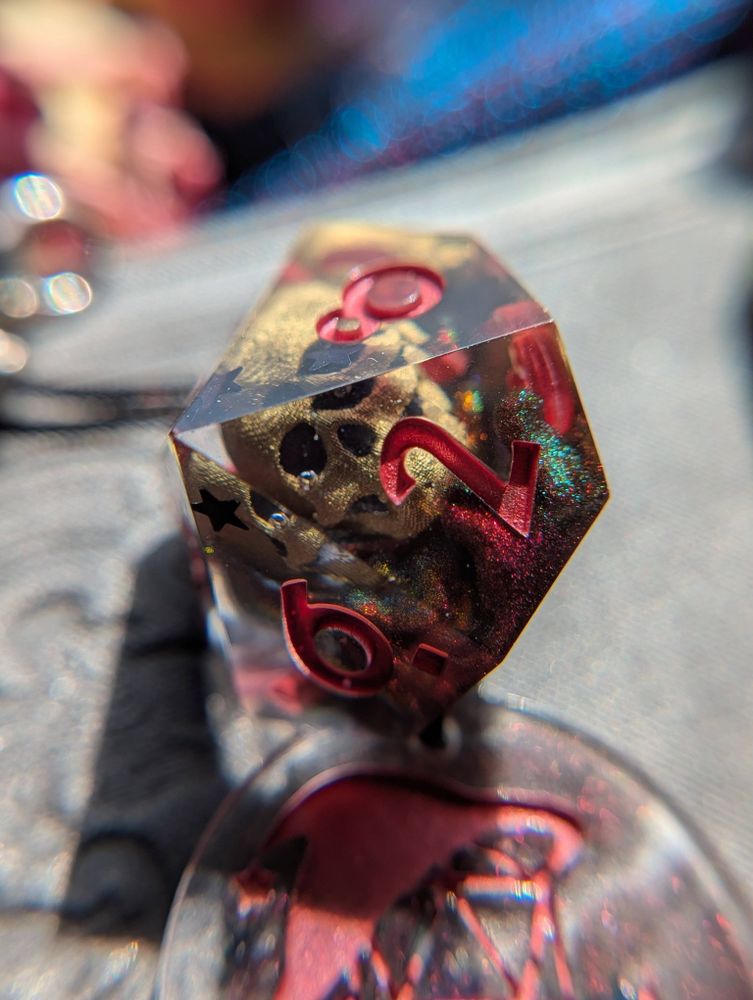Handmade dice featuring gold 3D printed skulls and swirls of red/green/yellow chameleon pigment, as well as black holographic stars.