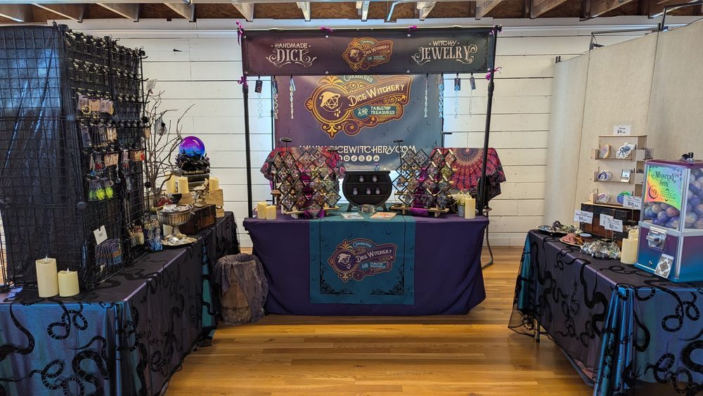 Dice Witchery vendor booth. The tables are covered in purple and blue cloths with black snakes. There are displays with jewelry, handmade dice sets, stickers, and a mystery spin gashapon machine.