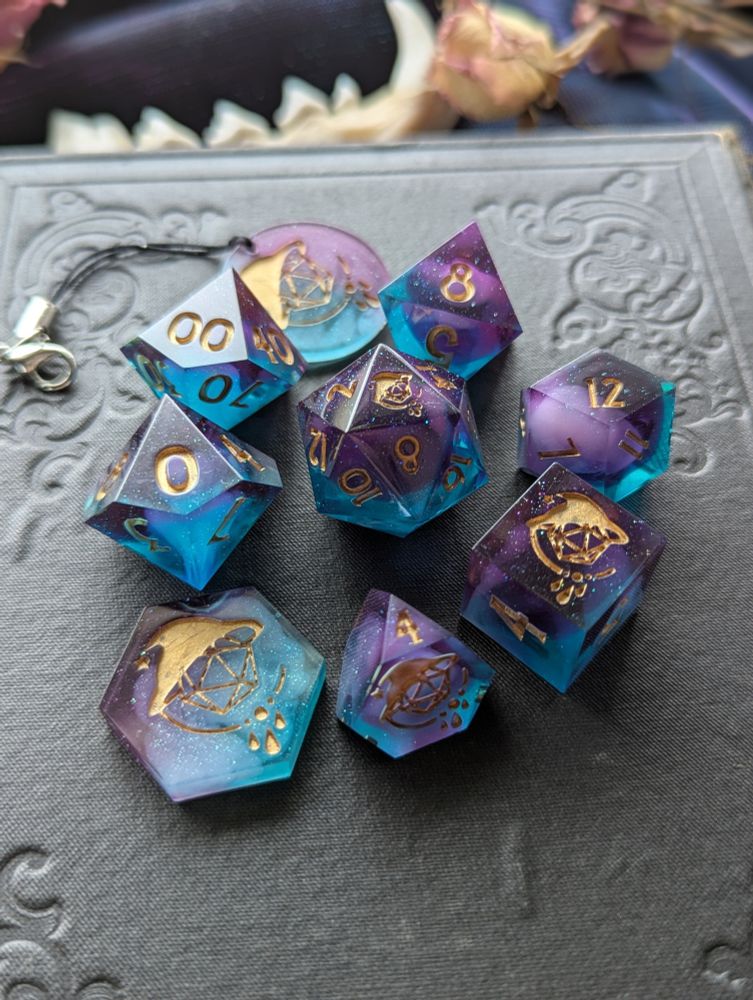 Handmade dice with white swirls in purple and teal transparent resin. They are inked gold.