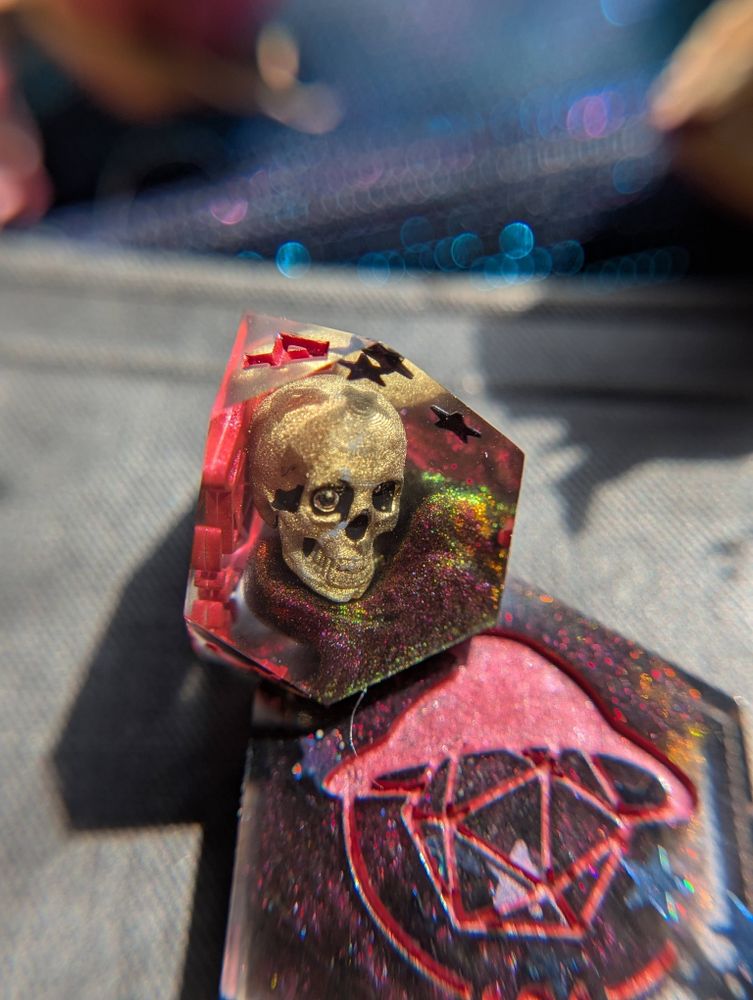 Handmade dice featuring gold 3D printed skulls and swirls of red/green/yellow chameleon pigment, as well as black holographic stars.
