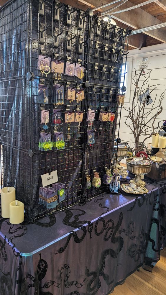 Dice Witchery vendor display with a grid wall of jewelry and pins. The tablecloth is a blue/purple metallic with black snakes.
